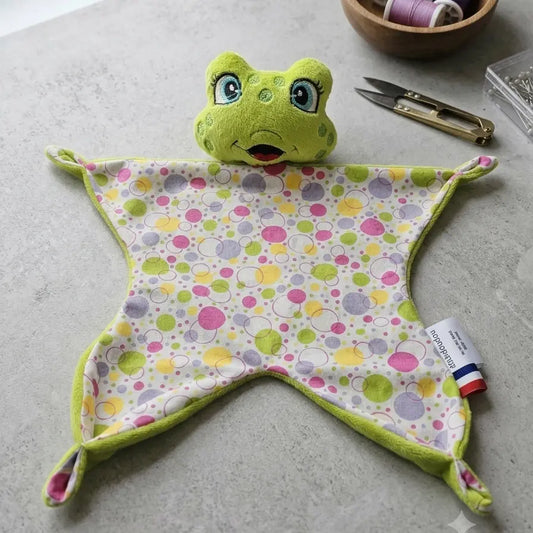 Doudou plat grenouille verte à motifs pois multicolores posé sur un plan de travail